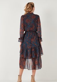 Pitkä, läpikuultava mekko, jossa on paisley-kuvio tummansinisissä ja ruosteenvärisissä sävyissä. Mekossa on kerroksellinen helma, joustava vyötärö ja nappahihansuut.