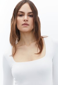 Wit shirt met lange mouwen, met een vierkante halslijn en een strakke, aansluitende pasvorm. De stof heeft een zachte, rekbare textuur en een minimalistische uitstraling.
