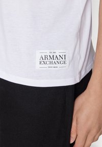 Biała bawełniana koszulka z czarną metką Armani Exchange naszytą na dolnym lewym boku. Koszulka ma gładką fakturę i luźny fason.