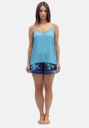 Vrouw staat blootsvoets, draagt een lichtblauwe mouwloze top en marineblauwe korte broek met een zeeschildpadprint op een effen achtergrond.