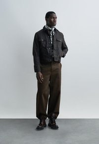 Bărbat purtând o jachetă scurtă de culoare închisă, eșarfă cu model, pantaloni largi maro și cizme închise cu șireturi, stând în picioare pe un fundal simplu și deschis la culoare.