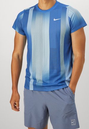Mężczyzna w niebieskiej koszulce Nike z krótkim rękawem i wzorem oraz w jasnoniebieskich spodenkach Nike, z ręką w kieszeni, stoi na jednolitym tle.