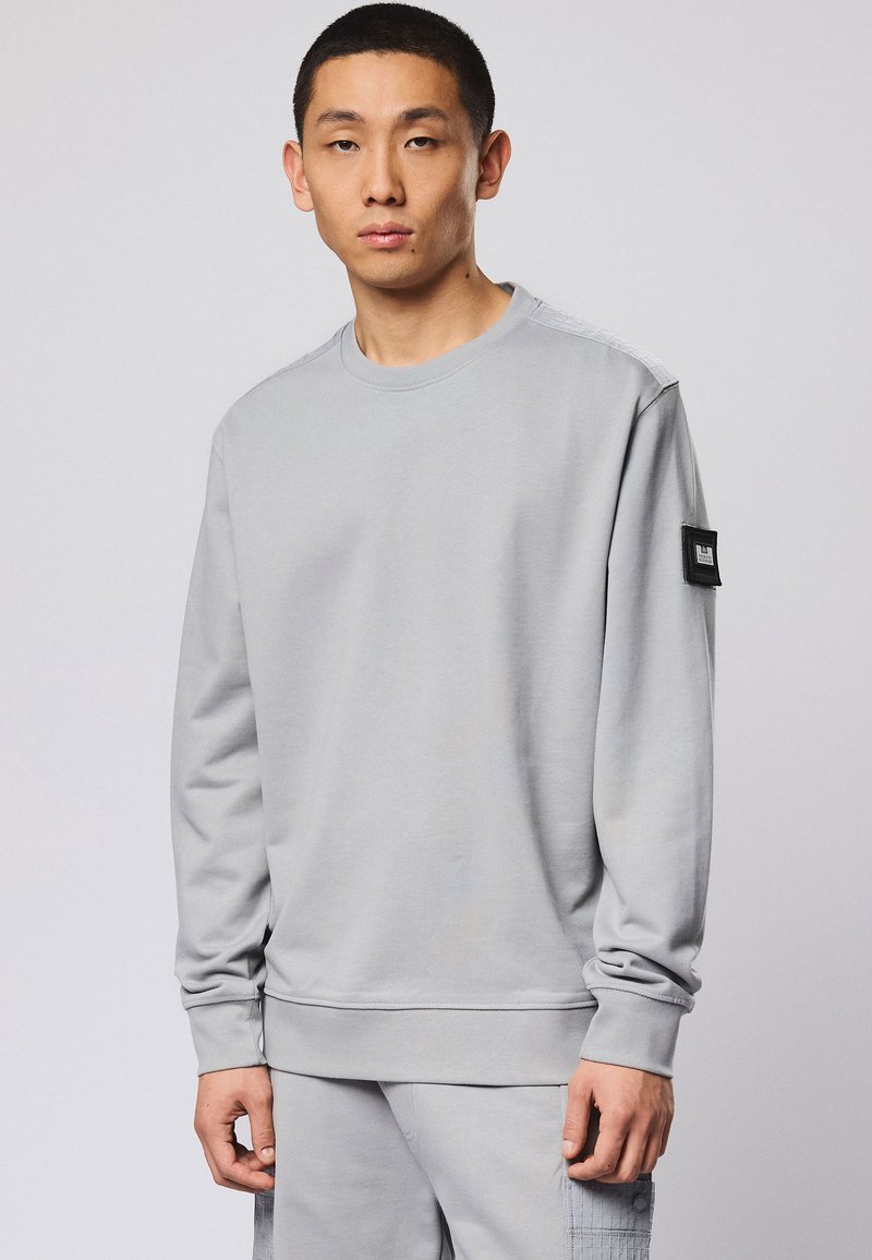Homme portant un sweat-shirt à manches longues gris clair avec un patch noir sur la manche gauche, assorti à un pantalon gris clair.