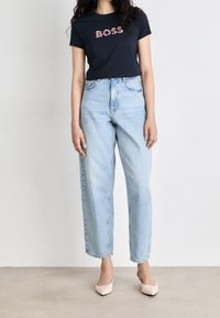 Žena oblečená v námořnickém modrém tričku s nápisem "BOSS" ve květinových písmenech, světlých modrých džínách s vysokým pasem a béžových špičatých balerínách.