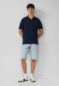 Marineblauwe polo met korte mouwen en een zak, gedragen met lichte denim shorts. Witte sneakers met zwarte accenten. Eenvoudig en casual ontwerp.