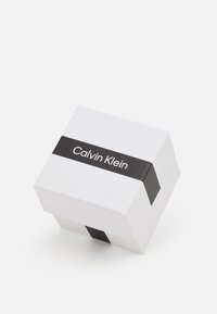 Fehér ajándékdoboz fekete szalaggal, amelyen a "Calvin Klein" felirat szerepel. A doboz négyzet alakú, sima tapintású és letisztult dizájnú.