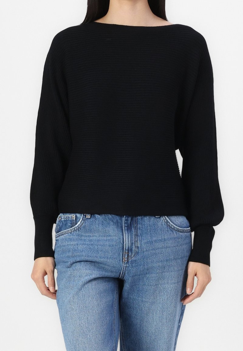 Femme portant un pull noir à manches longues côtelé avec des manches ballon et un jean bleu taille haute sur un fond blanc.