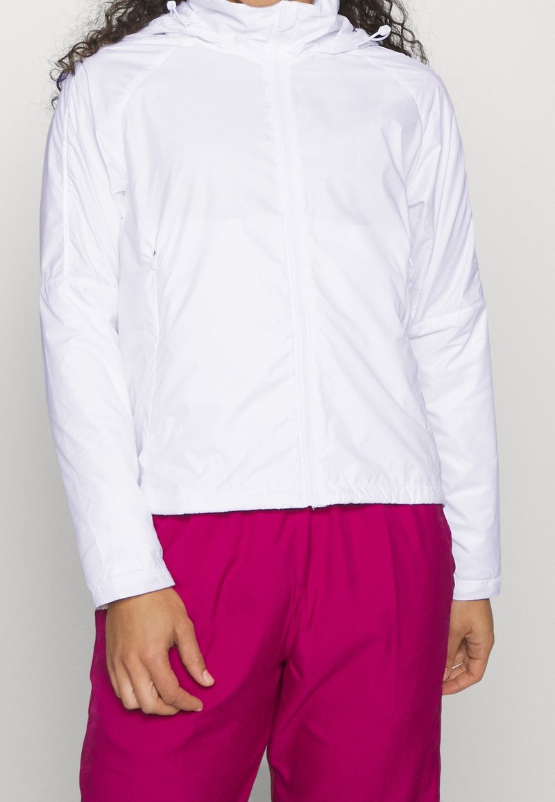 Personne portant une veste blanche zippée avec capuche et un pantalon rose vif, debout devant un fond clair uni.