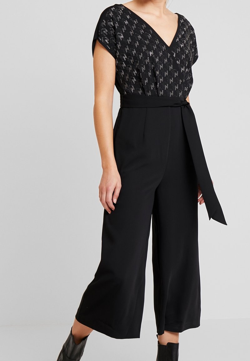 Femme portant une combinaison noire avec un haut à col en V à motifs, un pantalon à jambes larges et une ceinture nouée à la taille, associée à des bottines noires.