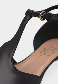 Sandale plate en cuir noir avec bride en T et petite boucle dorée sur une semelle intérieure beige avec texte de la marque.