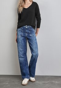 Svart V-ringad tröja, blå vida jeans med framfickor, blekt tvätt, och vita sneakers med beige detaljer på en grå bakgrund.