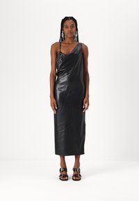 Svart skinnkjole med en skulderdesign, med en teksturert bodice. Modellen har på seg svarte sandaler med spenner, stående mot en hvit bakgrunn.