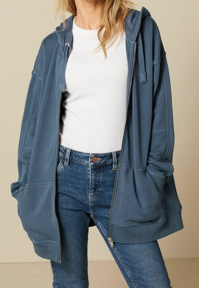 Femme portant un t-shirt blanc côtelé rentré dans un jean bleu avec un sweat à capuche bleu ample et dézippé avec poches avant et cordons.