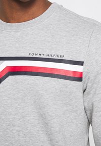 Nærbillede af en grå crew neck skjorte med diagonale striber i sort, hvid og rød, samt teksten "Tommy Hilfiger" ovenfor striberne.