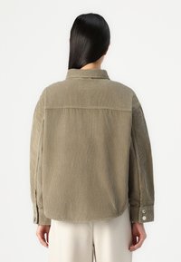 Veste en velours côtelé vert olive avec une coupe décontractée. Elle possède des coutures d'épaule, des poignets boutonnés et une finition lisse et texturée.