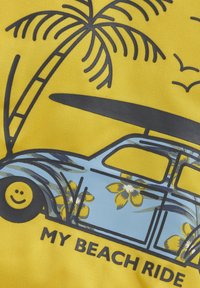 Gul tyg med en grafisk bild av en ljusblå vintagebil med blommiga detaljer, en surfbräda på taket och palmträdsmotiv. Texten nedanför lyder "MIN STRANDBIL."