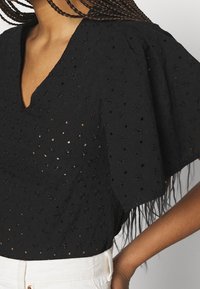 Černá halenka s výstřihem do V, krátkými nabíranými rukávy a květinovými průstřihy; v kombinaci s bílými kalhotami s vysokým pasem.