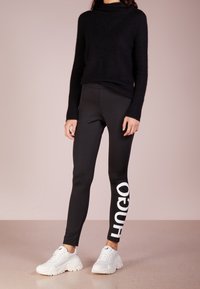 Mustapaita, jossa on pehmeä, teksturoitu pinta, yhdistettynä kiiltäviin mustiin legginsseihin, joissa on suuri valkoinen logo. Täydennä ulkoasu valkoisilla lenkkareilla.