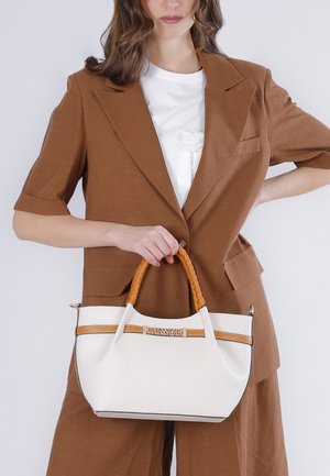 Donna con completo marrone che tiene una borsa bianca con manici intrecciati color cuoio e logo dorato del marchio "CAENORI" sul davanti.