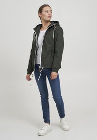 Mørk grønn lett jakke med hette, hvit glidelås og snorer. Har to frontlommer og ribbestrikkede mansjetter. Kombinert med blå jeans og hvite sneakers.