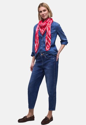 Vrouw draagt een blauw denim overhemd en spijkerbroek met een rood patroon sjaal en bruine loafers, staand met één hand in de zak.