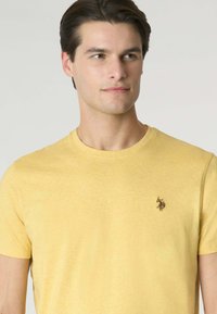 T-shirt gialla a maniche corte in tessuto morbido con scollatura rotonda e un piccolo logo marrone a sinistra del petto. Design semplice e classico.