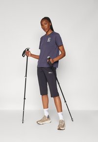 Mørkeblå t-skjorte med korte ermer og mønstret lomme, kombinert med mørke, avkuttede bukser og turstaver. Modellen har på seg beige sneakers og hvite sokker.