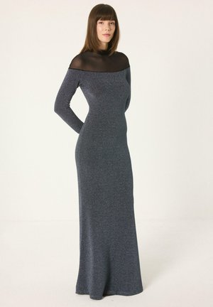 Femme debout portant une longue robe ajustée gris foncé avec un col noir transparent et des manches longues sur un fond clair uni.