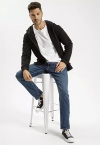 Czarna hoodie na zamek z białą koszulą, w połączeniu z niebieskimi dżinsami i czarnymi trampkami z białą podeszwą, siedzący na białym stołku.