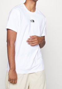 Camiseta blanca de algodón con un pequeño logotipo negro en el pecho. Diseño de cuello redondo estándar con mangas cortas y un ajuste relajado.
