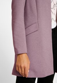 Gros plan sur la main d'une personne à côté d'une veste texturée mauve avec un rabat de poche plié, un pantalon noir et une chemise à motifs partiellement visible.