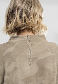 Beige genser med strukturert strikk, subtilt camouflasjemønster og glidelås bak med metallhardware.