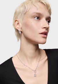 Collana in argento con anelli ovali, dotata di una chiusura decorativa e piccoli ciondoli. Il modello indossa un top nero e orecchini abbinati.