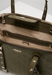 Olivengrøn totebag med præget logo mønster, guldfarvet hardware, lynlås lukning og glat indvendig foret. Har en lynlåslomme.