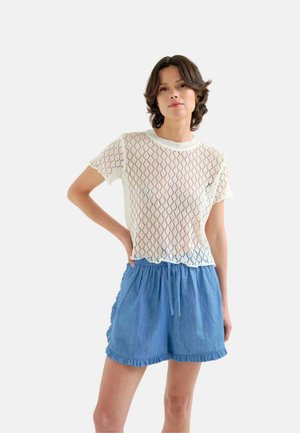 Mujer con una camiseta corta de encaje blanca y pantalones cortos azules con cintura elástica y volantes, de pie con una mano en la cadera.