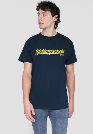 Junger Mann mit lockigem Haar, der ein dunkelblaues "Yellowjackets"-T-Shirt und eine blaue Jeans trägt, und nach links blickt vor einem schlichten Hintergrund.