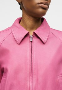 Κοντινό πλάνο ενός ατόμου που φοράει ένα ροζ δερμάτινο μπουφάν με μυτερό γιακά και φερμουάρ μπροστά, εμφανίζοντας μέρος της γνάθου και του λαιμού του.