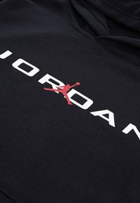 Crna dukserica s glatkom teksturom koja ima istaknuti bijeli natpis "JORDAN" i crvenu siluetu loga, centriranu u području prsnog koša.