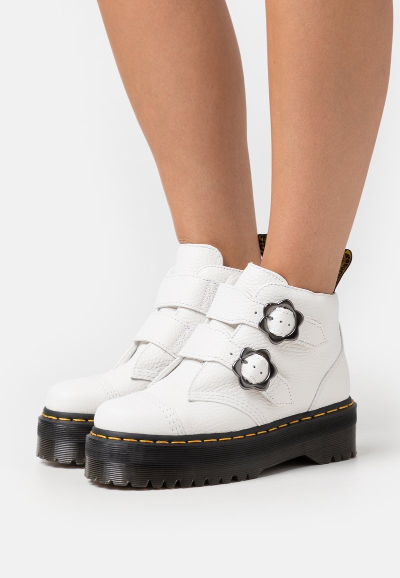 Bottes courtes en cuir blanc avec semelles épaisses noires, coutures jaunes, deux boucles en forme de fleur, portées sur des jambes nues sur un fond blanc.