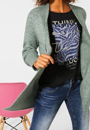Personne portant un cardigan vert par-dessus un t-shirt noir imprimé et un jean bleu, debout près d'une chaise rose sur un fond blanc.