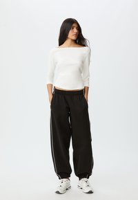 Witte aangesloten top met driekwart mouwen gecombineerd met zwarte, loszittende broek met witte zijstrepen en elastische boorden. Witte sneakers.