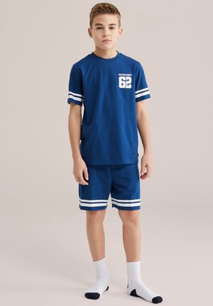 Junge steht, trägt ein blaues T-Shirt mit dem Schriftzug "Established 62" und passende blaue Shorts, weiße Socken mit schwarzen Zehen und Fersen.