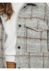 Grå og brun ternet uldskjortejakke med en frontlomme og sorte knapper, lagd ovenpå en hvid ribbet sweater.