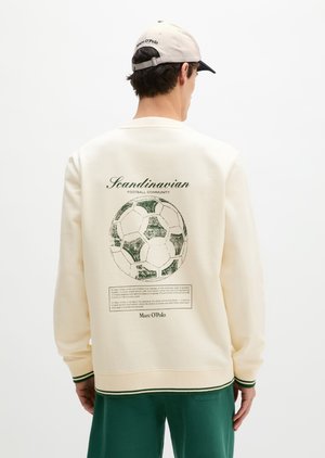 Mann iført kremfarget genser med grønn vintage fotballgrafikk og teksten "Scandinavian Football Community" på ryggen, kombinert med grønne bukser.