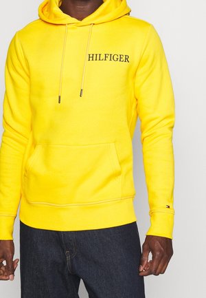 Mężczyzna w jaskrawożółtej bluzie z kapturem z napisem "HILFIGER" na piersi oraz małym logo na rękawie, w połączeniu z ciemnoniebieskimi dżinsami.