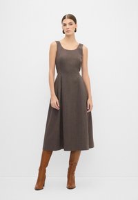 Robe midi sans manches marron avec un corsage ajusté et une jupe évasée, réalisée dans un tissu texturé. Portée avec des bottes à talons hauts marron.