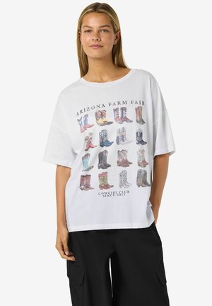 Ung kvinde iført hvid oversized t-shirt med farverige cowboystøvler print og teksten "Arizona Farm Fair" samt sorte cargobukser.