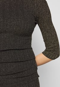 Svart ribbad topp med trekvartslånga ärmar, med metalliska guldprickar och lager på lager ruffle-detaljer vid midjan. Stretchtyg.