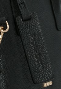 Dettaglio di una borsa a mano in pelle nera con un'etichetta testurizzata con la scritta "PICARD" in rilievo, accenti in hardware dorato e uno sfondo liscio.