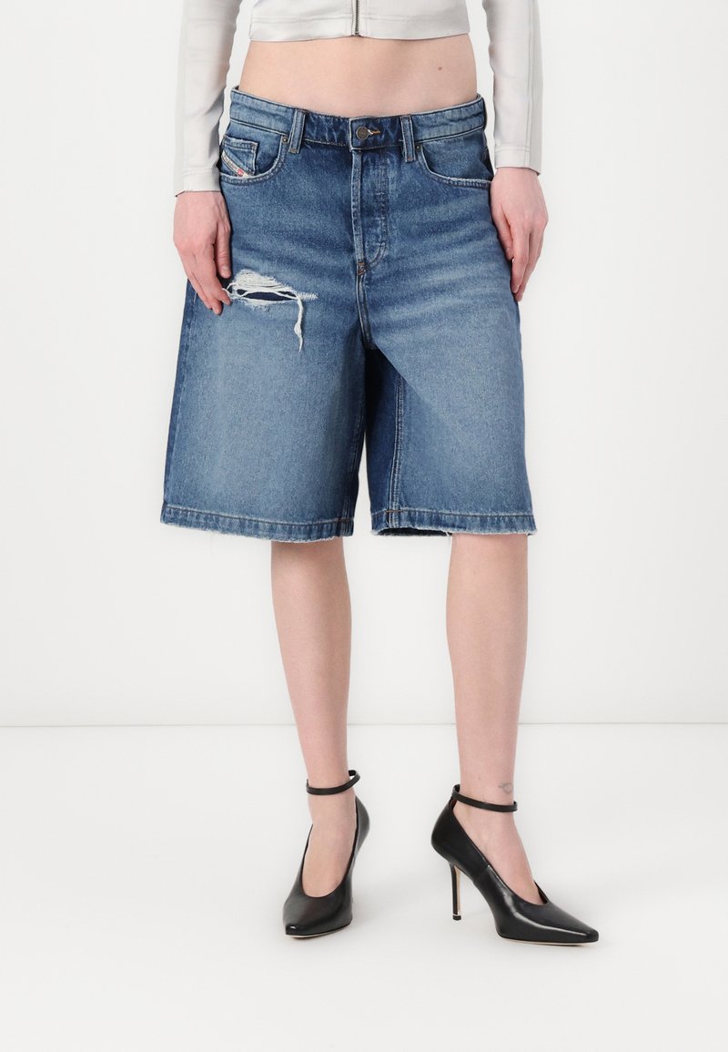 Shorts denim berwarna biru sedang dengan potongan longgar, memiliki bagian depan yang robek dan hem yang digulung. Dipadukan dengan sepatu hak tinggi berwarna hitam.